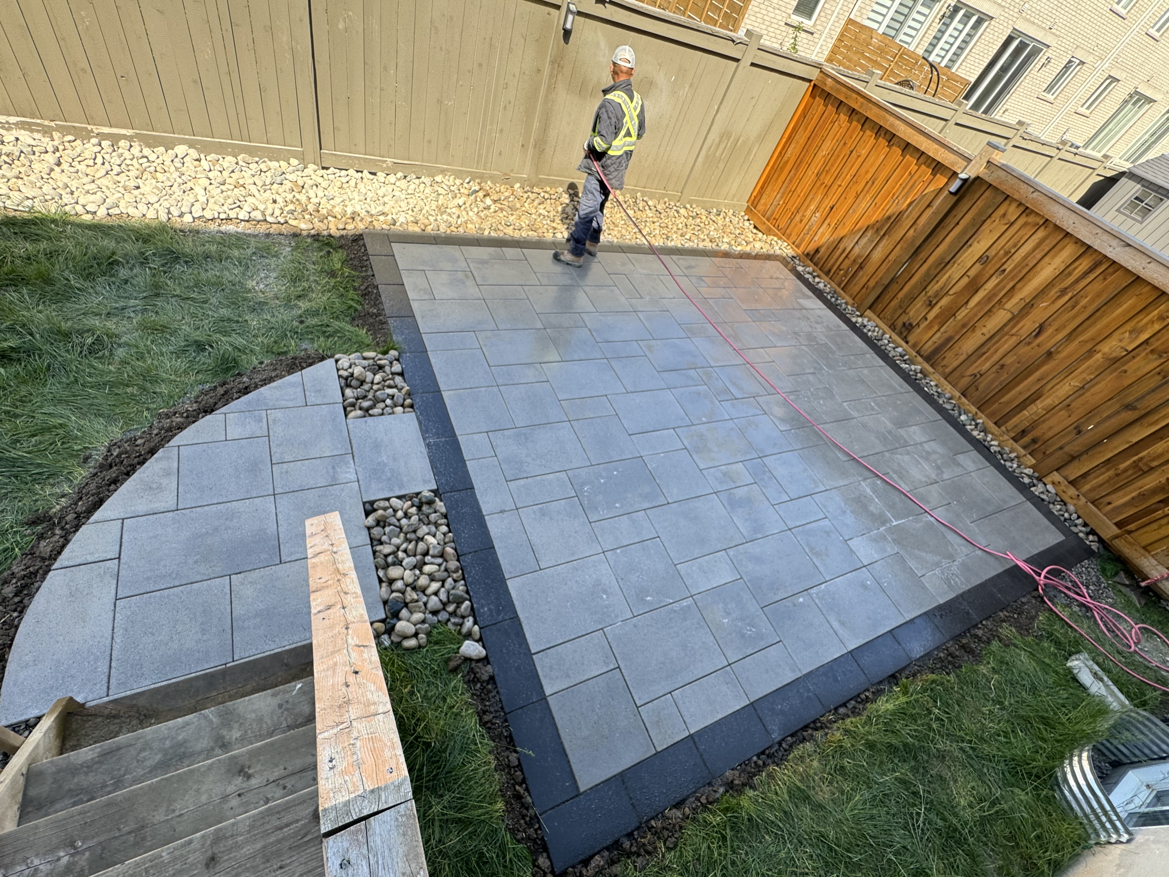 Worker finishing interlock backyard patio with curved pathway and river rock accents