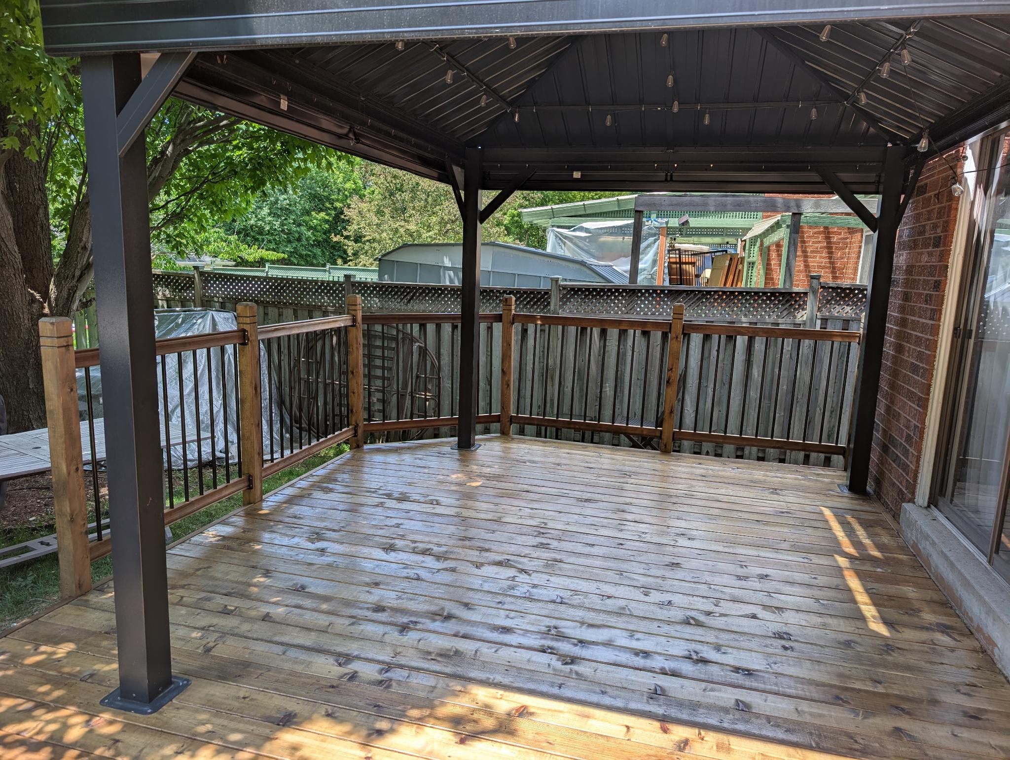 Deck under metal gazebo before staining — fresh unstained boards, dark metal posts